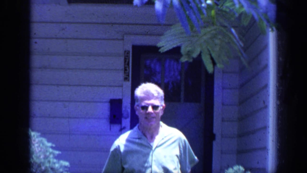 Man in sunglasses walks forward from a doorway framed by foliage.