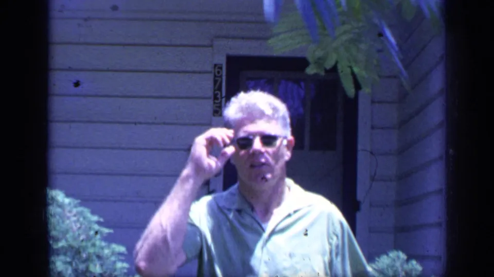 The man raises a hand to his sunglasses near the doorway of a Florida house in blue-shifted 8mm footage.