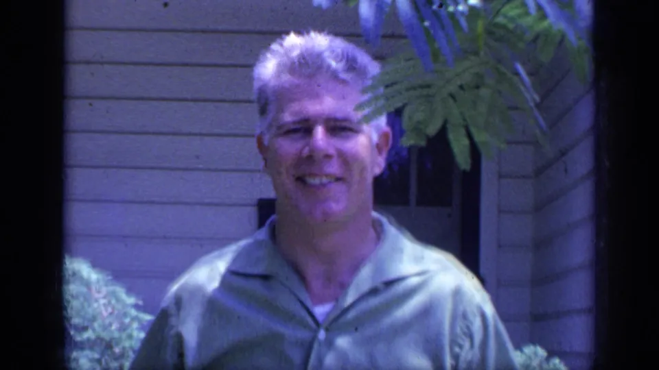 A smiling man in a short-sleeve shirt faces the camera, with a house doorway and foliage behind him in 1968 Florida footage.