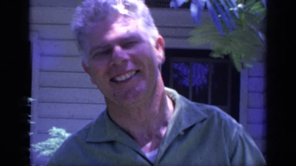 1968 Florida home-movie frame of a smiling adult counselor in a green collared shirt outside a light-sided building.