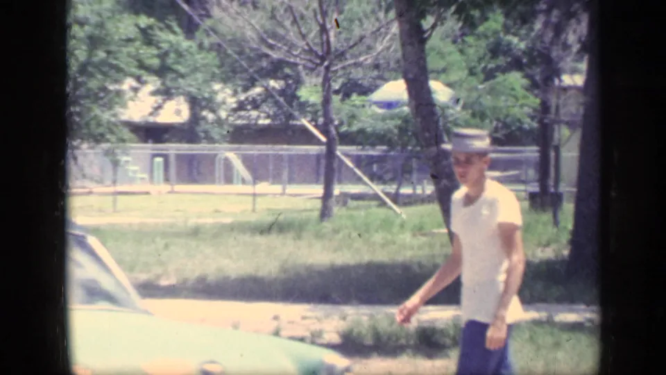 A man in warm-weather clothing walks beside a pale parked car, with trees and camp buildings behind him in 1968 Florida footage.