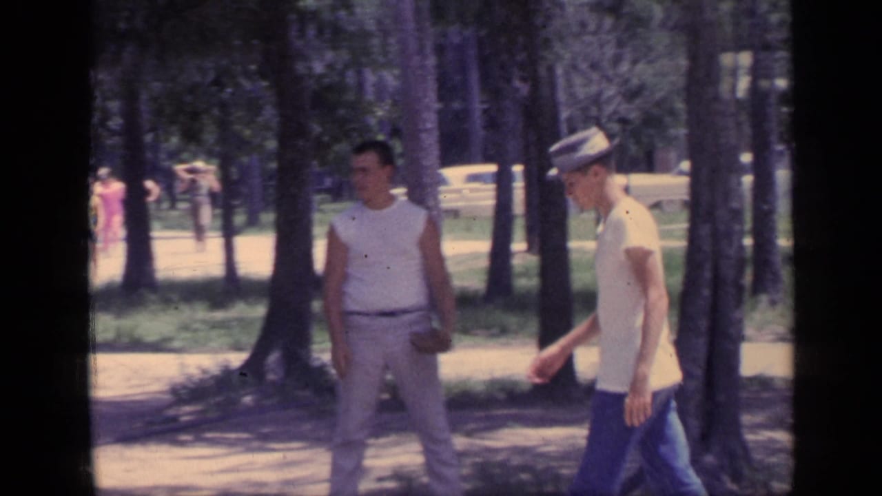 A young man in a hat walks past another young man holding a baseball glove in a shaded Florida park, with paths and trees in the background.