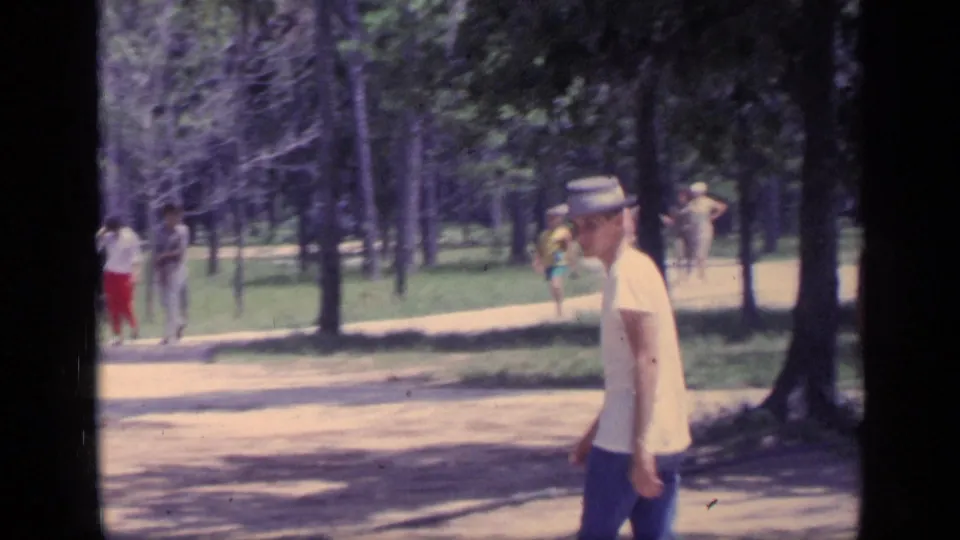 The hat-wearing young man continues across the frame in a 1968 Florida park scene, with distant walkers and shaded trees visible behind him.