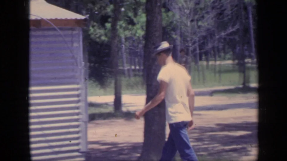 A young man in a brimmed hat walks beside a small metal-sided structure in the Florida park, with trees and a path in the background.