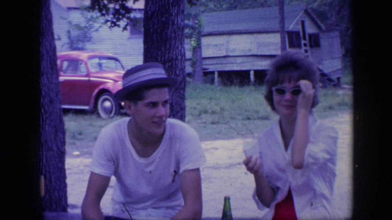 Young man in a hat sits beside young woman in sunglasses outdoors near trees and a red car.