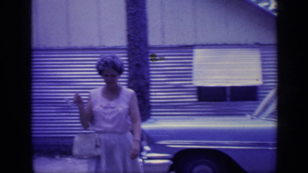 A woman in a light dress walks toward the camera in 1968 Florida footage, with a corrugated-sided building and parked car behind her.