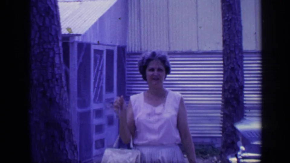The woman continues toward the camera in the Florida outdoor scene, framed by a tree trunk, screen door area, and corrugated building wall.