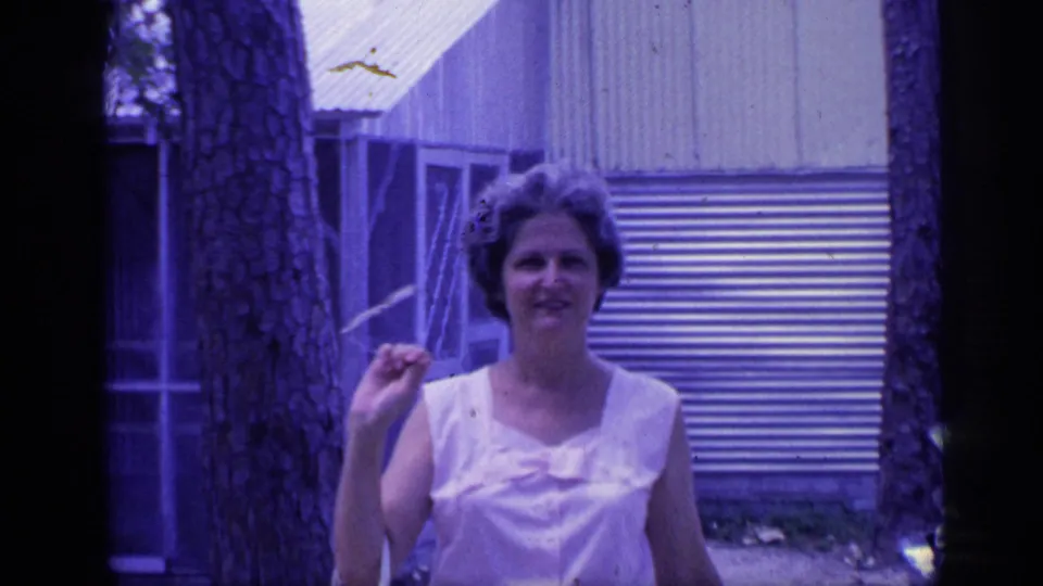 A woman smiles toward the camera in the 1968 Florida home movie, standing near a tree with corrugated siding and a screened doorway behind her.