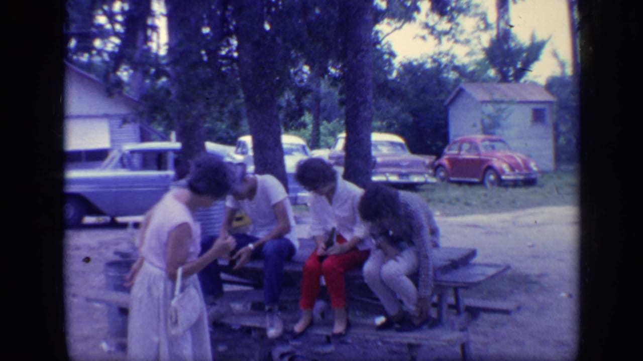Four adults gathered at a picnic table with parked cars and trees behind them.