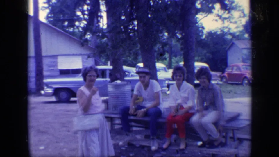 1968 Florida 8mm frame of four adults seated at a picnic table near a cylindrical trash can, with classic cars and trees behind them.