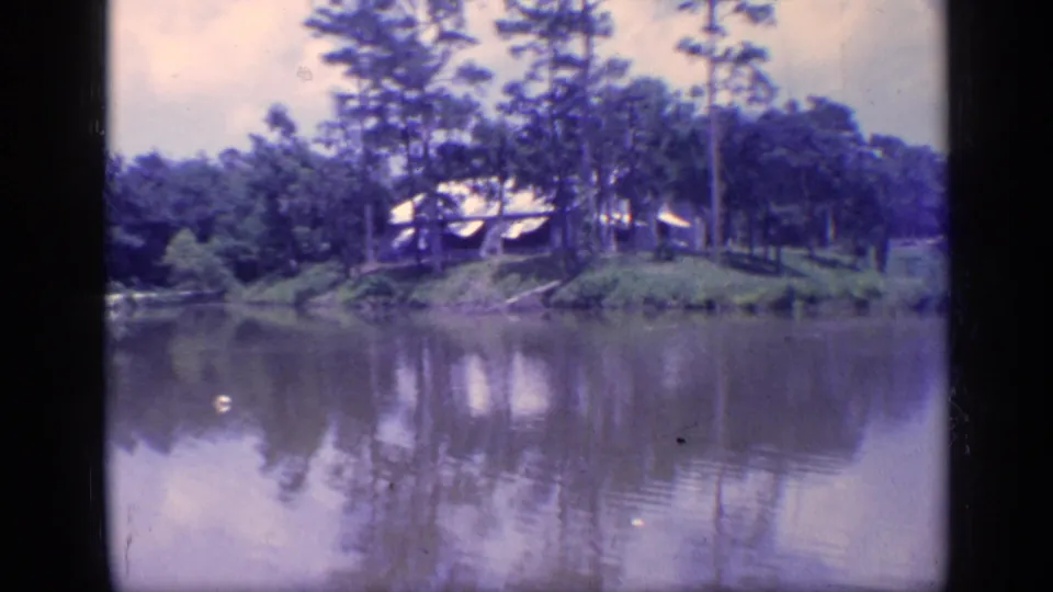 1968 Florida 8mm frame of a lakeside cabin set among pine trees, reflected in the pond water.