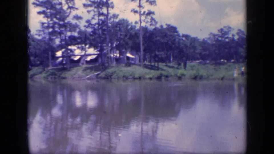 1968 Florida home-movie frame with a cabin on a pine-lined pond bank and muted reflections in the water.