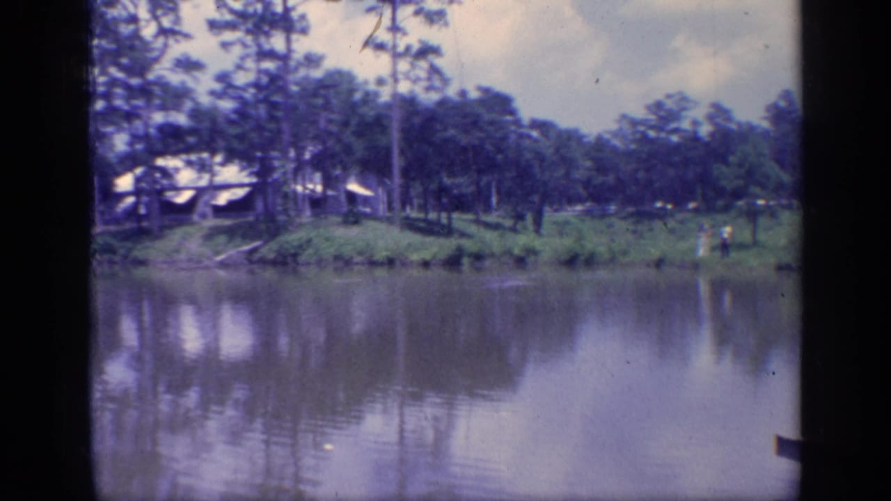 Calm water facing a tree-lined bank with small distant figures and structures among pines.