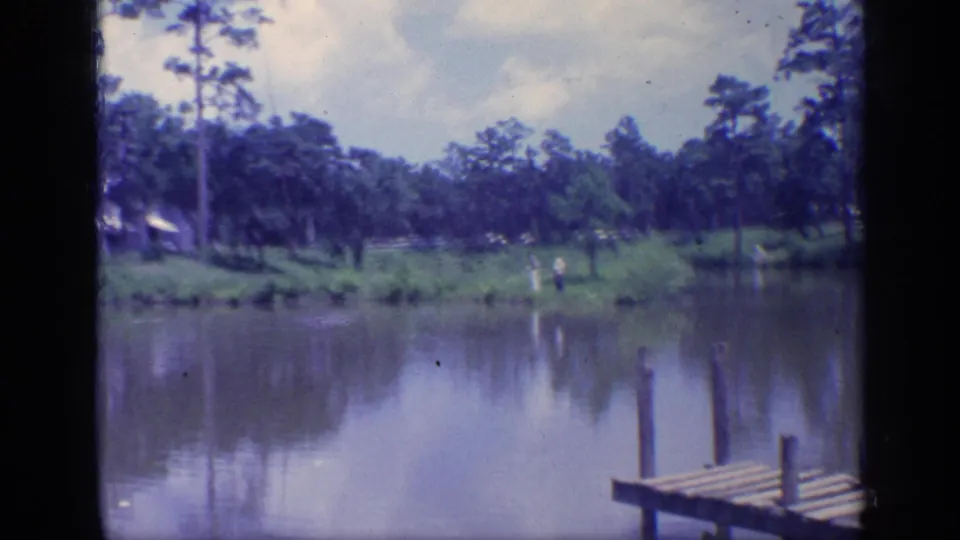 1968 Florida 8mm frame showing a wooden dock foreground, calm water, pine trees, and small distant figures on the far bank.