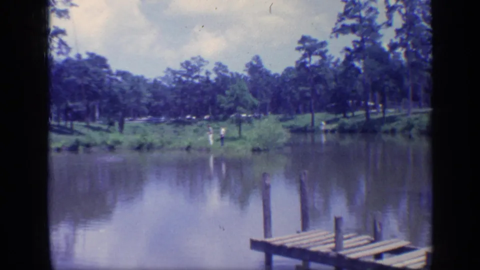 1968 Florida home-movie frame of a wooden pier, still muddy water, grassy bank, pine trees, and distant people on shore.