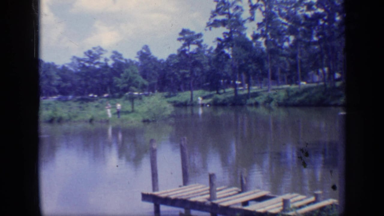 1968 Florida home-movie frame with a weathered dock, still pond water, pine trees, shrubs, and small people near the far bank.