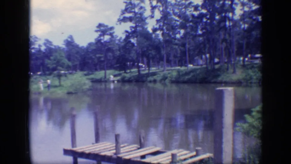 1968 Florida 8mm view of a wooded pond with dock posts in the foreground, a grassy bank, and distant people near the water.