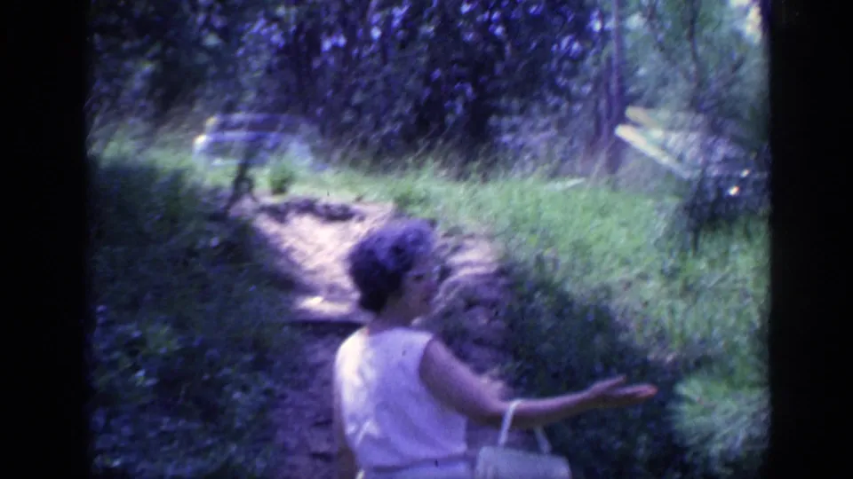1968 Florida 8mm frame of a woman in a light dress with a purse walking on a wooded dirt path.