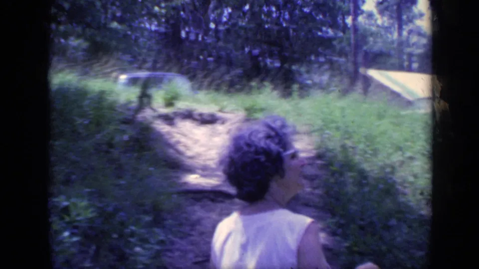 Seen from behind, the woman continues along the dirt path through grass and trees in the supplied Florida 1968 home-movie setting.