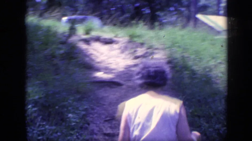 The woman walks away along a narrow dirt path, with grass, trees, and a pale background structure visible in the 1968 Florida footage.