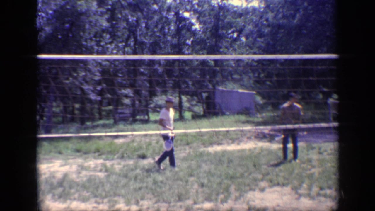 Two men stand on opposite sides of a badminton net in a grassy wooded yard.