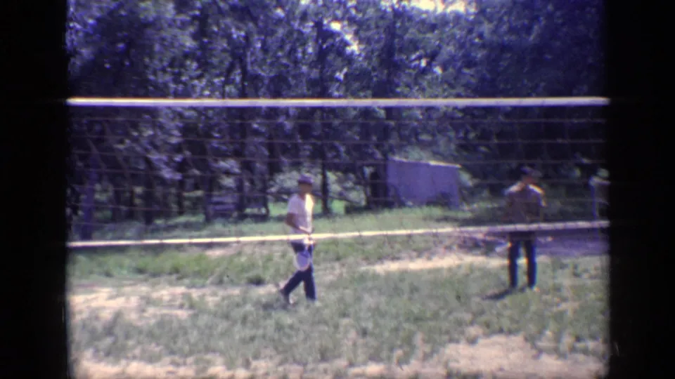 1968 Florida home-movie frame showing two young men with rackets near a portable badminton net in a sunlit wooded yard.