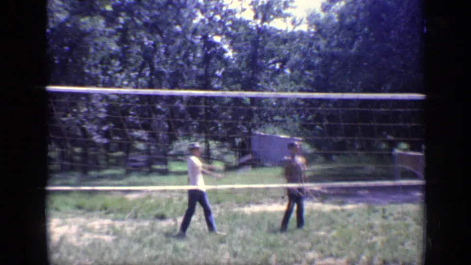 A soft 8mm frame shows the two badminton players moving through a rally in a sunlit yard with trees and a small structure behind them.