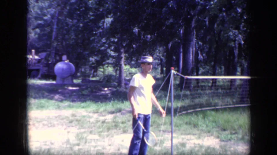 One badminton player in a light shirt and hat stands closer to the camera beside the net, with trees and yard objects in the background.
