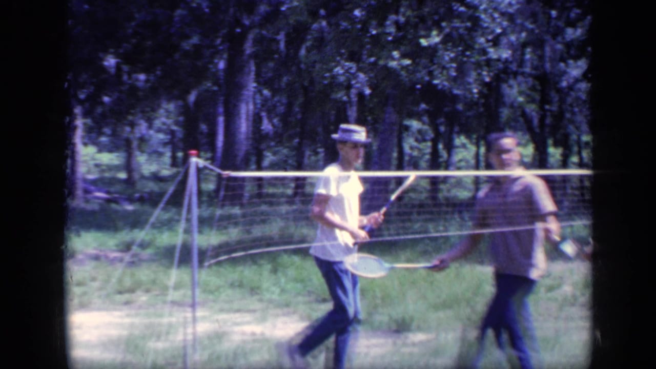 Two men play badminton across a backyard net near trees.