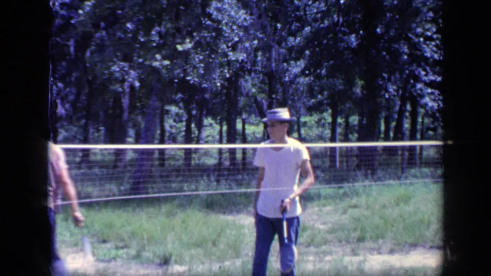 The 1968 Florida home movie shows a man in a white T-shirt and hat standing beside a backyard badminton net with trees behind him.