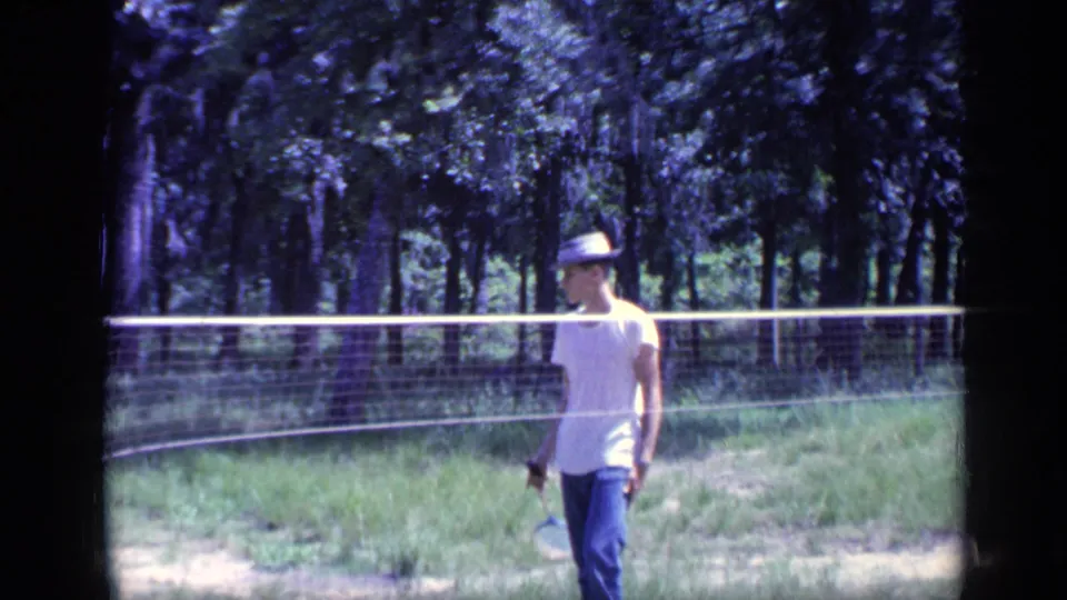 A man in a white T-shirt and brimmed hat walks along the backyard badminton court in 1968 Florida 8mm footage.