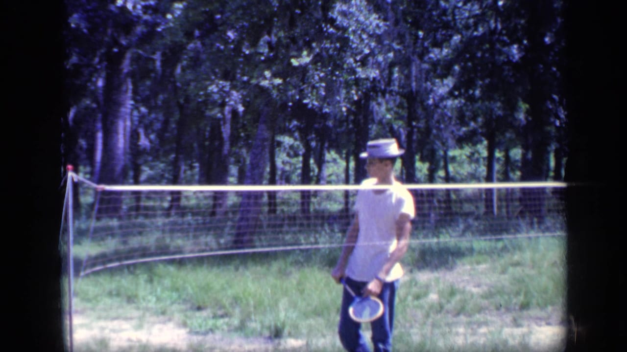 Young man holding a badminton racket near a net in a wooded grassy clearing.