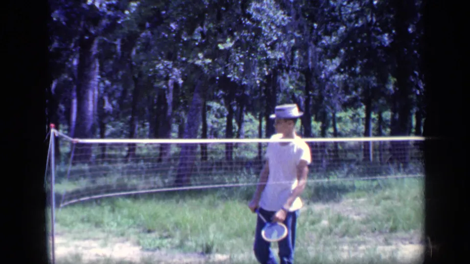 1968 Florida 8mm frame showing a young man in a white T-shirt and hat holding a badminton racket beside a net outdoors.