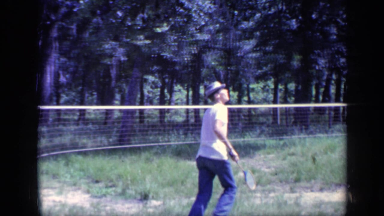 Man with a badminton racket walks along a net with trees behind him.