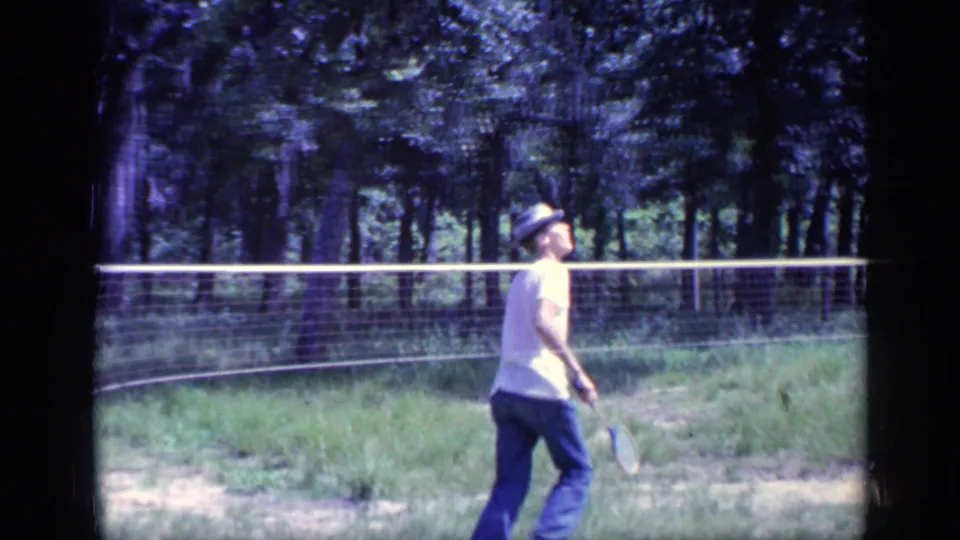 1968 Florida home-movie frame of a man in casual clothes moving beside a badminton net in a tree-lined outdoor area.