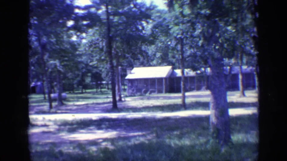 1968 Florida 8mm frame showing rustic campground cabins beyond trees, grass, and a dirt path.