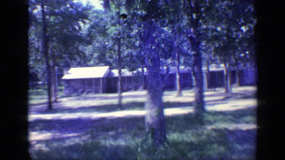 1968 Florida home-movie frame of campground cabins behind trees, with sunlit grass and a dirt track.