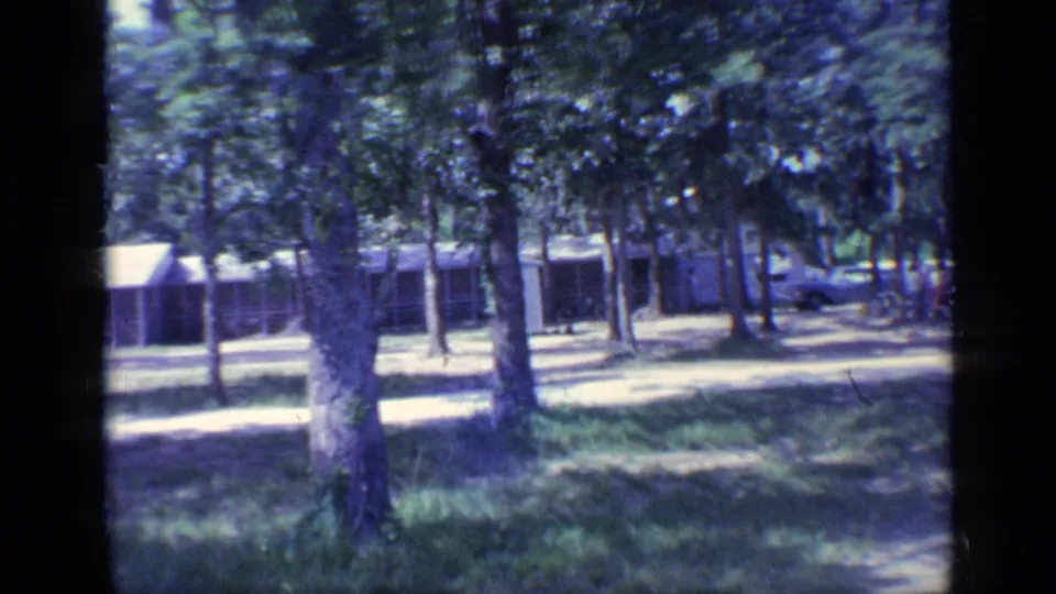 1968 Florida 8mm frame with cabins partly hidden by trees and a parked car in the background.