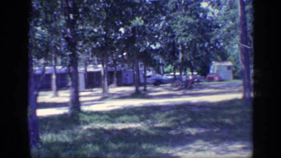 1968 Florida campground frame showing cabins, trees, dirt ground, and distant classic cars.