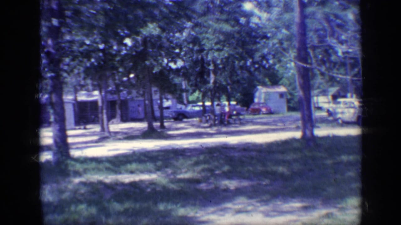 Shaded campground with trees, parked cars, a dirt path, and distant people near a small building.