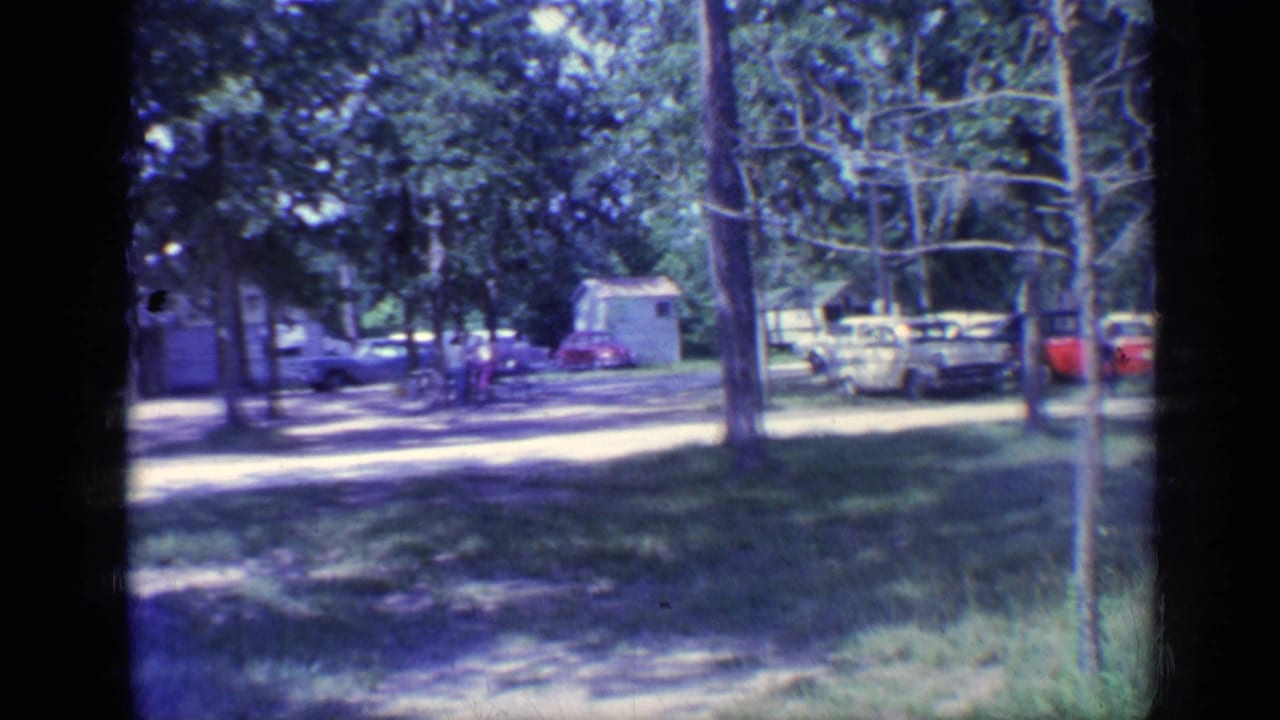 Wooded campground area with several parked cars, grass, trees, and people near a white building.