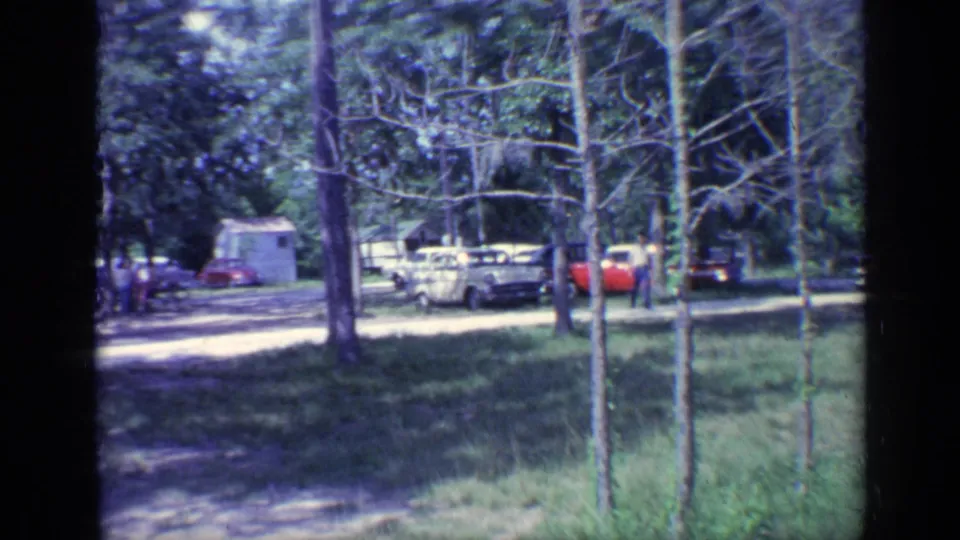 Foreground saplings partially frame a 1968 Florida campground with parked cars, grass, and a small white building beyond.