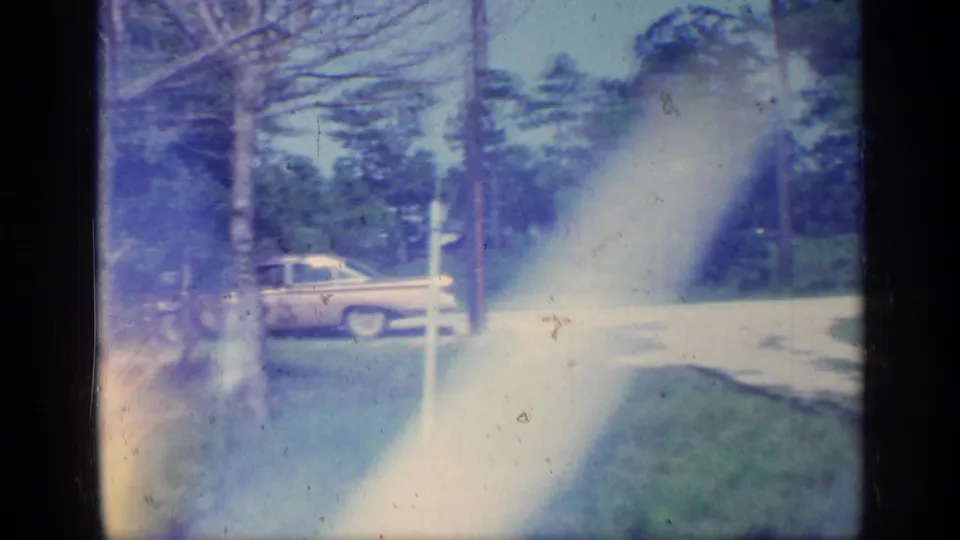 1968 Florida roadside frame with a pale car, trees, grass, and a broad blue-white exposure flare from the scanned 8mm film.