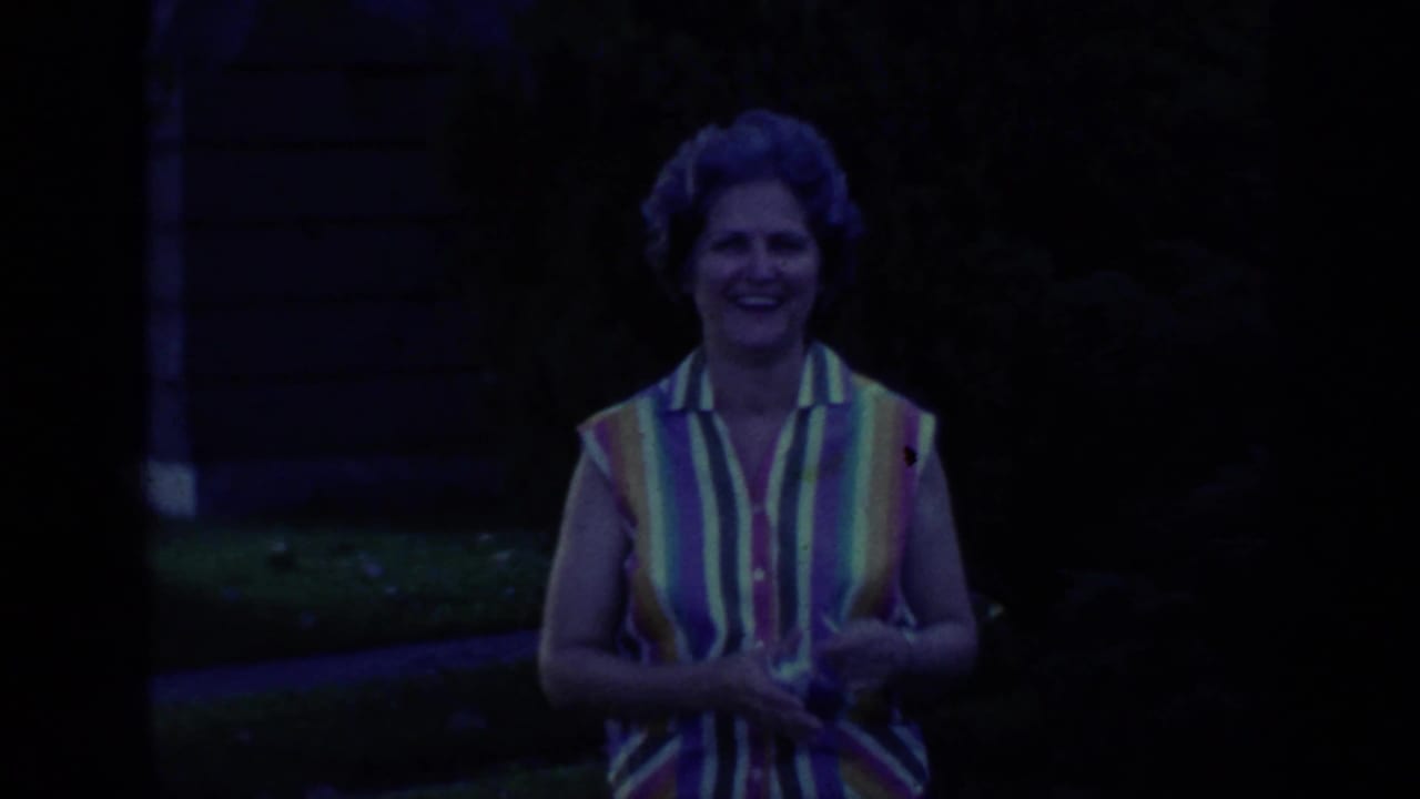 Dim frame of a smiling woman in a striped sleeveless blouse standing in a yard.