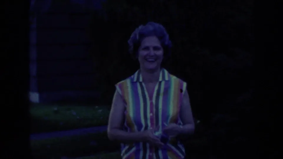 In a dark 1968 Florida 8mm frame, a smiling woman in a multicolored striped sleeveless blouse stands before lawn and shrubs.