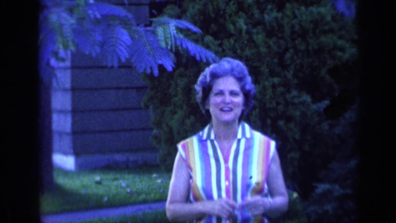 A smiling woman in a bright striped blouse is framed from the waist up in a Florida yard, with house siding and shrubs behind her.