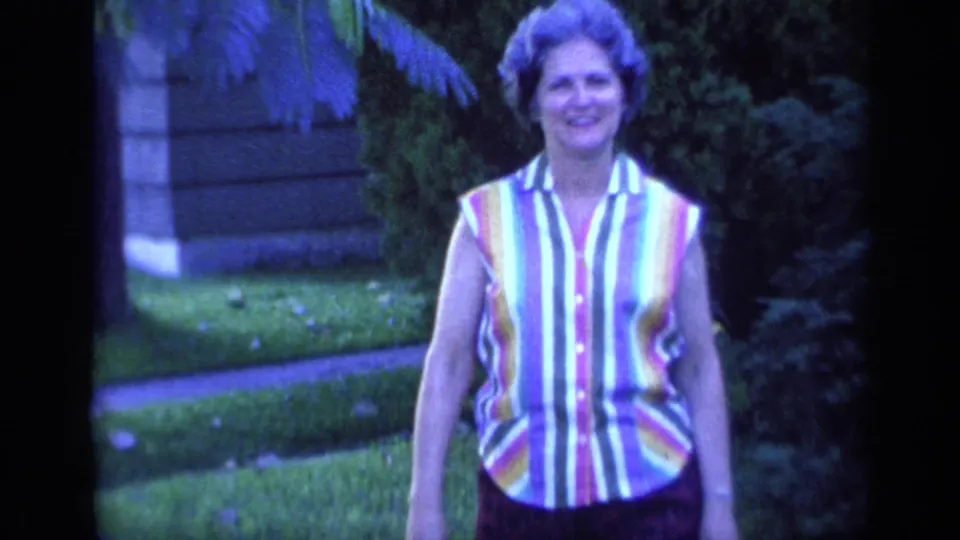 The woman walks closer to the camera in the 1968 Florida yard, wearing a striped sleeveless blouse with shrubs and a walkway behind her.