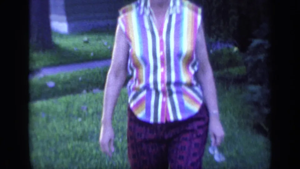A closer 1968 Florida home-movie frame shows the woman's striped blouse and purple patterned pants as she walks toward the camera.
