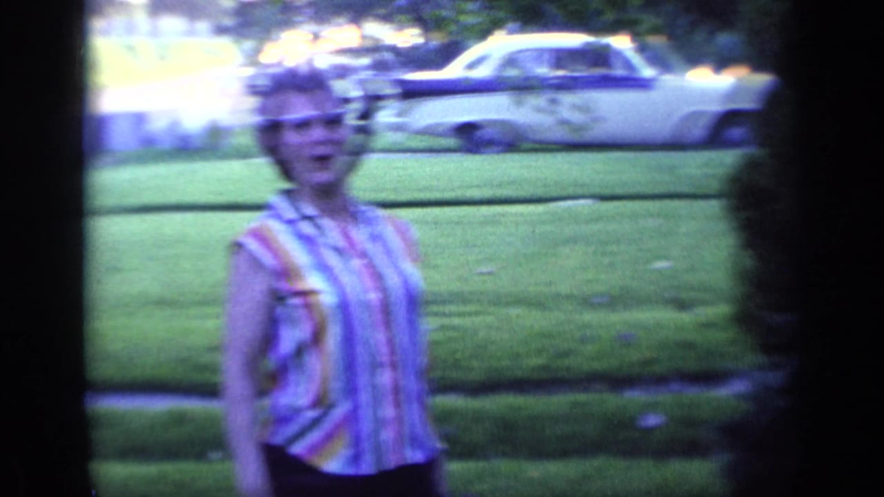 Woman with glasses stands on a lawn with a parked car behind her.