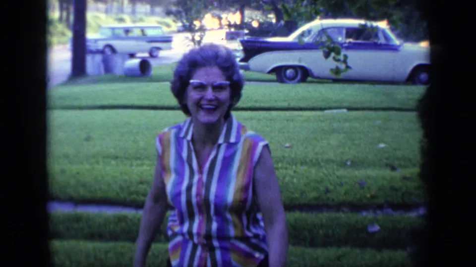 Smiling woman wearing sunglasses faces the camera on a suburban Florida lawn, with a parked classic car across the street.